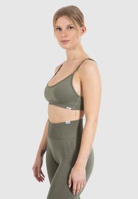 Sujeta sin costuras en color verde oliva con tirantes delgados, con costuras visibles y etiqueta de la marca en la parte delantera. Leggings de yoga en un color texturizado a juego.
