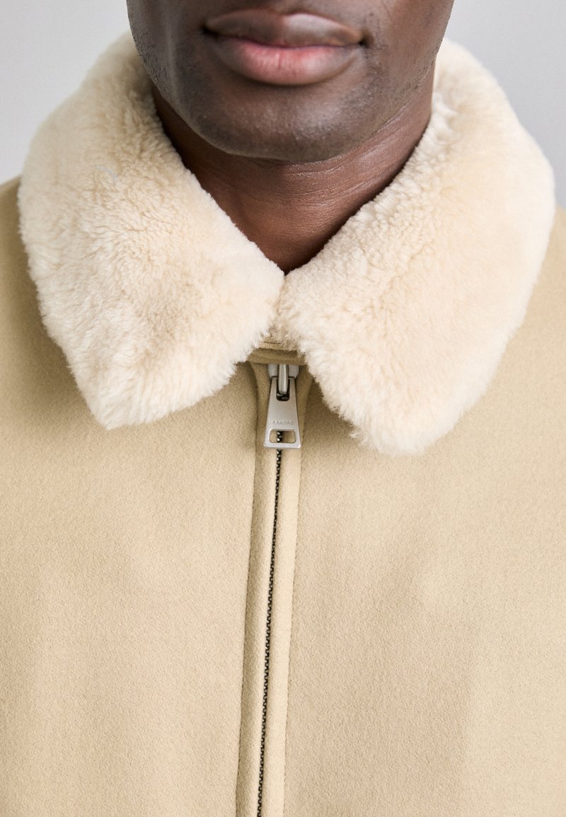 Beige jasje met een zachte, crèmekleurige nep-bont kraag. De voorkant heeft een zilveren ritsopening en een gladde, matte textuur.