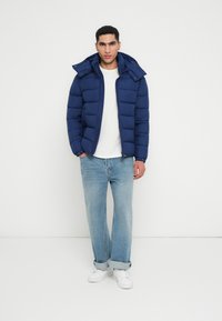 Marineblå pufferjakke med hætte, lynlås og ribbede manchetter. Sættes sammen med lyseblå afslappede jeans og hvide sneakers, stående mod en hvid baggrund.