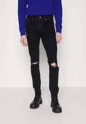 Persoon die zwarte gerafelde skinny jeans met kniegaten, zwarte veterlaarzen en een blauw shirt met lange mouwen draagt, staand op een effen achtergrond.