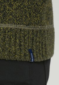 Groene en gele gebreide trui met een zichtbare zoom en een klein blauw label met de tekst "DENIM RIGHTS" bovenop zwarte broek.