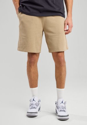 Beige cotton shorts with an elastic waistband and side pockets. Paired with white socks and white sneakers featuring gray accents.