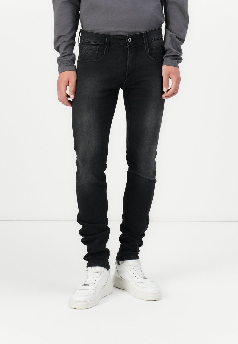 Personne portant un jean slim noir et des baskets montantes blanches, debout devant un fond blanc uni.