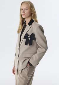 Beige op maat gemaakt blazer met zwarte revers, gestippelde textuur, en een decoratieve zwarte lintspeld met een zilveren accent.