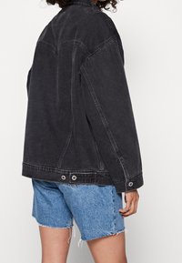 Giacca di jeans nera con una vestibilità comoda, cuciture visibili e due bottoni metallici all'orlo, abbinata a pantaloncini di jeans azzurri sfilacciati.