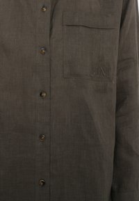 Camicia button-up marrone in lino con tasca e dettaglio ricamato "MM". Presenta bottoni scuri e un tessuto strutturato.