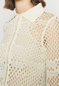 Helles beige Strickhemd mit einem strukturierten, offenen Maschenmuster, das über einen gerippten Kragen und eine Vorderknopfleiste verfügt.