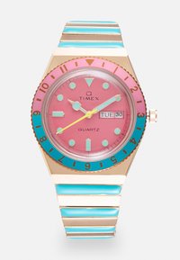 Timex Q kvarts kell roosa dialiga, türkiissiniste aktsentidega, kuldne korpus ja triibuline metalllink randmeese teal ja kuldse värviga. Omab päeva/kuupäeva näidikut.
