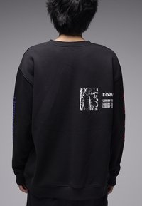 Μαύρο φούτερ με στρογγυλή λαιμόκοψη, με γραφικό σχέδιο στην πλάτη και χρωματιστά κείμενα σε κόκκινο και μπλε στα μανίκια.