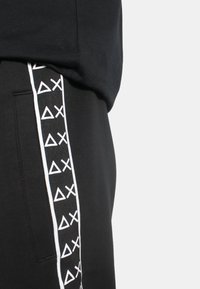 Pantaloni da tuta neri con nastro laterale bianco a motivo geometrico. Il tessuto sembra morbido e presenta un design con vestibilità rilassata.