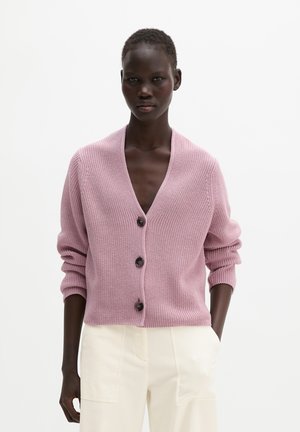 Personne à la peau foncée portant un cardigan léger en tricot rose clair boutonné et un pantalon blanc cassé, debout devant un fond blanc uni.