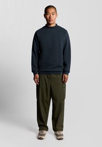 Donkerblauwe sweatshirt met een hoge hals, gecombineerd met olijfgroene cargobroeken met zijzakken. Gemaakt in combinatie met lichtgekleurde sneakers.