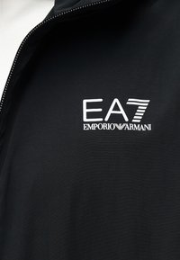 Μαύρο ένδυμα με λευκό λογότυπο EA7 Emporio Armani στο στήθος, με μερικώς ανοιχτό φερμουάρ ορατό στο πλάι.