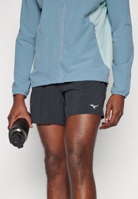 Blau und hellgrüne Sportjacke mit Reißverschluss und elastischen Bündchen, kombiniert mit schwarzen Shorts mit einem Logo. Die Person hält eine schwarze Wasserflasche.