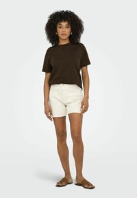 T-shirt in cotone marrone abbinato a pantaloni shorts in denim beige chiaro. Il modello indossa sandali a ciabatta, mettendo in risalto il design semplice e la vestibilità rilassata.