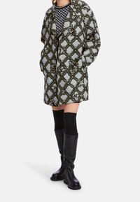 Cappotto a fantasia grigio chiaro con design esagonale verde scuro, caratterizzato da una vestibilità morbida e tasche. Stivali neri al ginocchio e calzini.