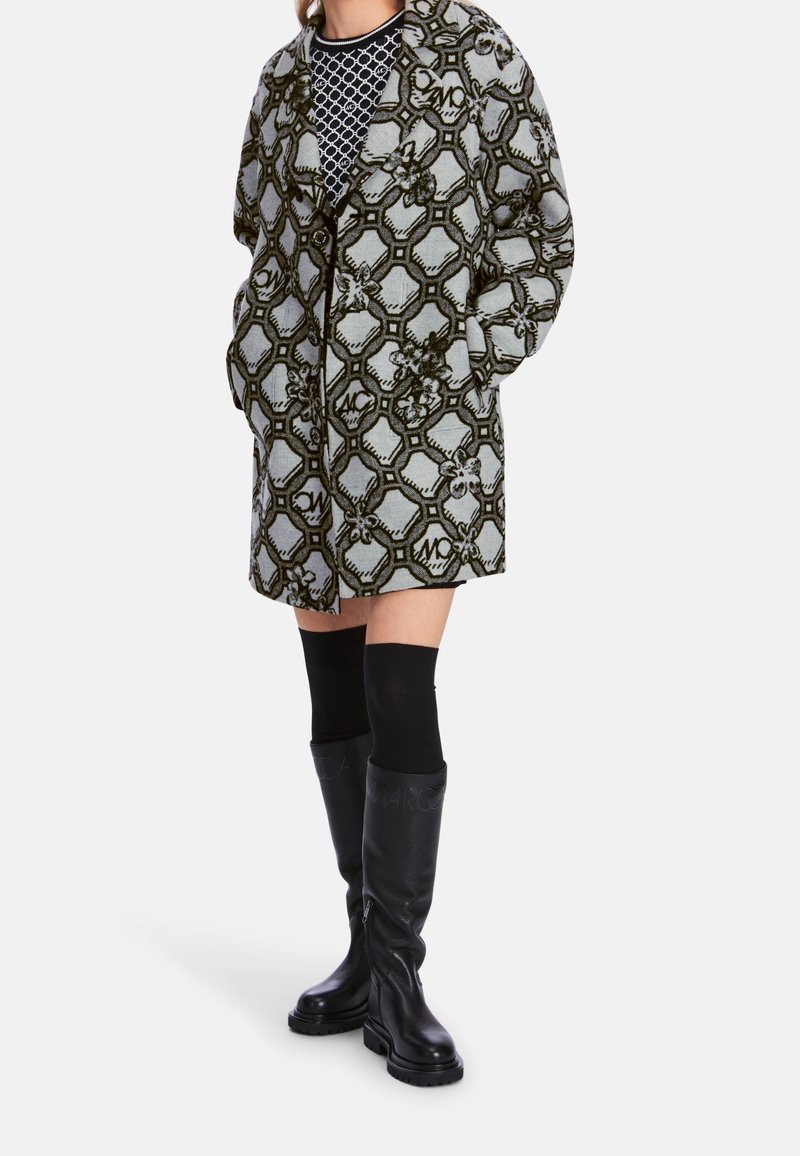 Cappotto a fantasia grigio chiaro con design esagonale verde scuro, caratterizzato da una vestibilità morbida e tasche. Stivali neri al ginocchio e calzini.