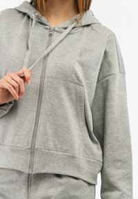 Graue Zip-Hoodie aus weichem Material. Mit einer Fronttasche, Kordelzügen und einem mit Kristallen verzierten Logo auf der Brust.