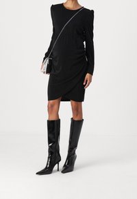 Robe noire à manches longues avec fronces à la taille, associée à des bottes noires à talons aiguilles montantes jusqu'au genou. Un petit sac bandoulière en argent est inclus.