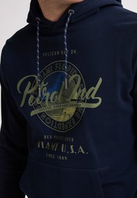 Felpa navy con stampa grafica frontale in verde, caratterizzata da testo e un design circolare. Include lacci e una tasca a marsupio.