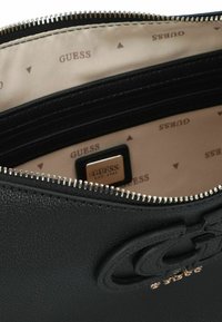 Sac à main en cuir noir avec un dessus zippé, une surface texturée, et un intérieur de marque en tissu beige imprimé du nom "GUESS".