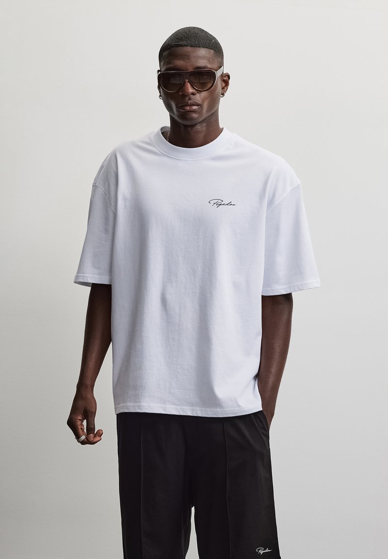 Άνδρας με oversized λευκό μπλουζάκι με μικρό λογότυπο στο στήθος, μαύρο παντελόνι, μεγάλα γυαλιά ηλίου και σκουλαρίκια, ποζάρει μπροστά σε απλό γκρι φόντο.