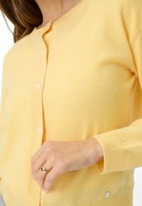 Gelber Strickcardigan mit rundem Ausschnitt, vorderen Knöpfen, gerippten Bündchen und einem kleinen Metall-Logoakzent nahe dem Saum.