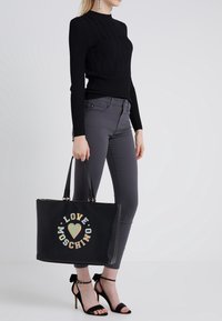 Zwarte lederen tote bag met gouden hardware, met het "LOVE MOSCHINO" logo in pasteltinten. Model draagt een zwart geribde trui en grijze broek.