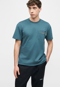 Νεαρός άνδρας με τιρκουάζ κοντομάνικο πουκάμισο με φερμουάρ στην τσέπη του στήθους και μαύρο παντελόνι, στέκεται με το ένα χέρι στην τσέπη, μπροστά σε απλό φόντο.