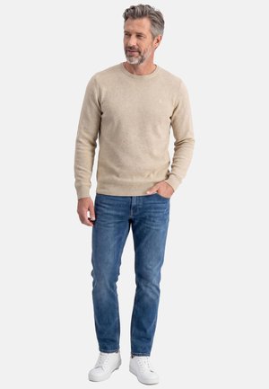 Uomo con capelli grigi e barba, che indossa un maglione beige, jeans blu e sneakers bianche, in piedi con una mano in tasca, guardando di lato.