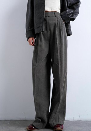 Personne portant un pantalon gris taille haute à jambes larges, un crop top blanc, une veste en cuir noire et des chaussures rouge foncé, debout contre un mur uni.