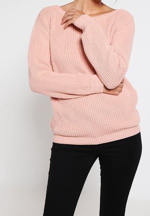 Personne portant un pull en maille épaisse rose clair à manches longues, associé à un pantalon noir ajusté, les bras croisés au niveau de la poitrine.