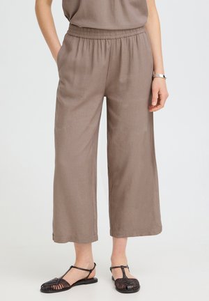 Kobieta ubrana w taupe szerokie spodnie 7/8 z elastycznym pasem oraz czarne plecione sandały na płaskiej podeszwie z paskiem w kształcie litery T na białym tle.