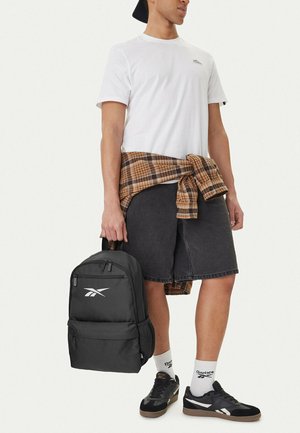 Junger Mann in weißem T-Shirt und schwarzer Shorts hält einen schwarzen Reebok-Rucksack, mit einem karierten Hemd um die Taille gebunden und passenden Reebok-Schuhen.