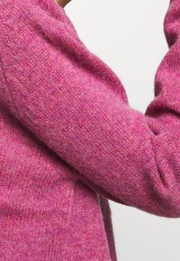 Rosa ulltröja med en texturerad stickning, med en något lös passform och långa ärmar. Färgen visar subtila variationer.