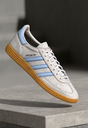 Baskets Adidas Spezial gris clair avec des bandes bleues et une semelle en gomme, montrées en suspension au-dessus d'une surface en béton.