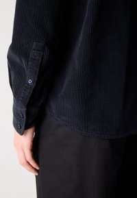 Camisa de pana azul marino con textura acanalada, con puños abotonados y un corte relajado. El fondo es neutro.