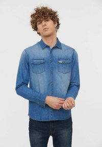 Chemise en denim bleu clair avec des manches longues, poches avant et fermeture à boutons. Présente une coupe régulière et une texture lisse.