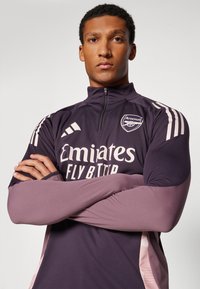 Lila Trainingsjacke mit hohem Kragen, ausgestattet mit hellrosa Akzenten, dem "Emirates"-Logo und einem Arsenal-Wappen auf der Brust.