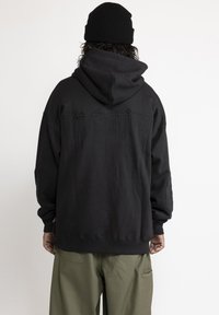 Zwarte hoodie met een getextureerd bergontwerp in zwarte draad op de achterkant. Oversized pasvorm, geribbelde manchetten en zoom, gedragen met groene broek.