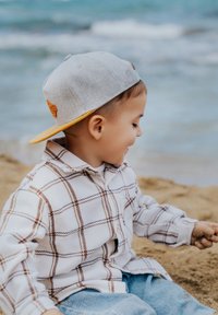 Casquette snapback grise avec accent orange ; chemise en flanelle à carreaux blancs et marron ; jeans bleu clair ; plage de sable en arrière-plan.
