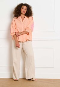 Žena stojaca so založenými rukami, oblečená v voľnej broskyňovej košeli s ružovými vzormi a širokých krémových nohaviciach proti bielej stene.