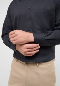 Camicia nera con bottoni e finitura testurizzata, con taschino sul petto e maniche lunghe, abbinata a pantaloni beige.