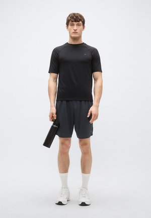 Black short-sleeve athletic shirt and dark gray shorts, holding a black water bottle. White sneakers with white ankle socks. Clean, minimal design.