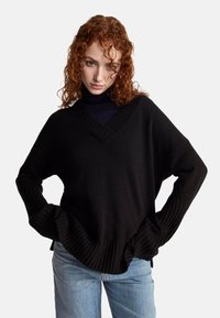 Czarny sweter z dzianiny z dekoltem w serek, ściągaczami przy mankietach i u dołu, luźny krój, zestawiony z jasnoniebieskimi dżinsami. Teksturowana, miękka tkanina.