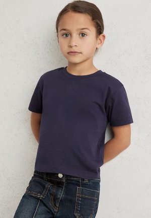 Ung pige med brunt hår står mod en lys tekstureret væg, iført en mørkeblå t-shirt og mørkeblå jeans med lommer.