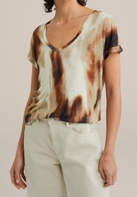 Top z krótkim rękawem, w dekolcie w serek, w kremowej tkaninie z brązowymi i czarnymi abstrakcyjnymi wzorami. Połączony z jasnymi spodniami.