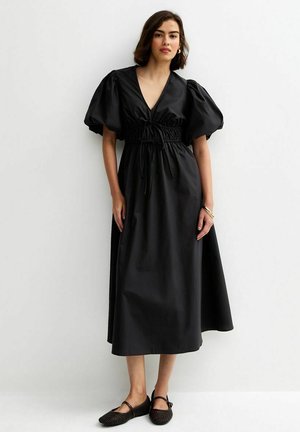 Kobieta w czarnej sukience midi z bufkami, dekoltem w kształcie litery V, taliowaną talią oraz czarnymi płaskimi butami, stojąca na tle jednolitej białej ściany.