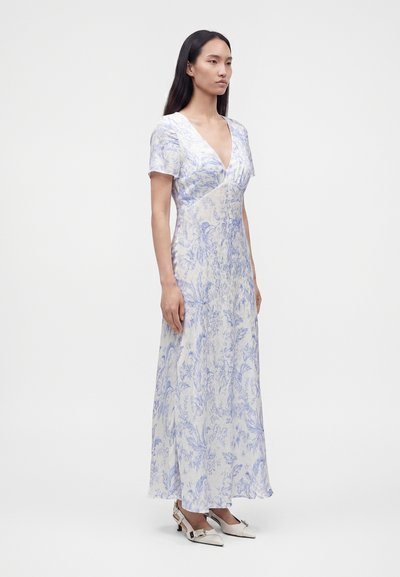 Femme portant une robe blanche longue jusqu'aux chevilles avec un imprimé floral bleu, manches courtes, un col en V profond et des chaussures blanches pointues avec des boucles.