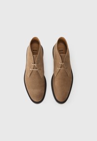 Un par de zapatos de ante marrón con cordones y suelas negras etiquetados como "BOSS PREMIUM COMFORT" sobre un fondo blanco liso.